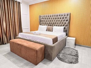 A bedroom with a tufted headboard, a large bed with white and beige bedding, a wooden accent wall, a grey furry rug, and a wooden bench. - THE NOOKHOUSE (Calabar)