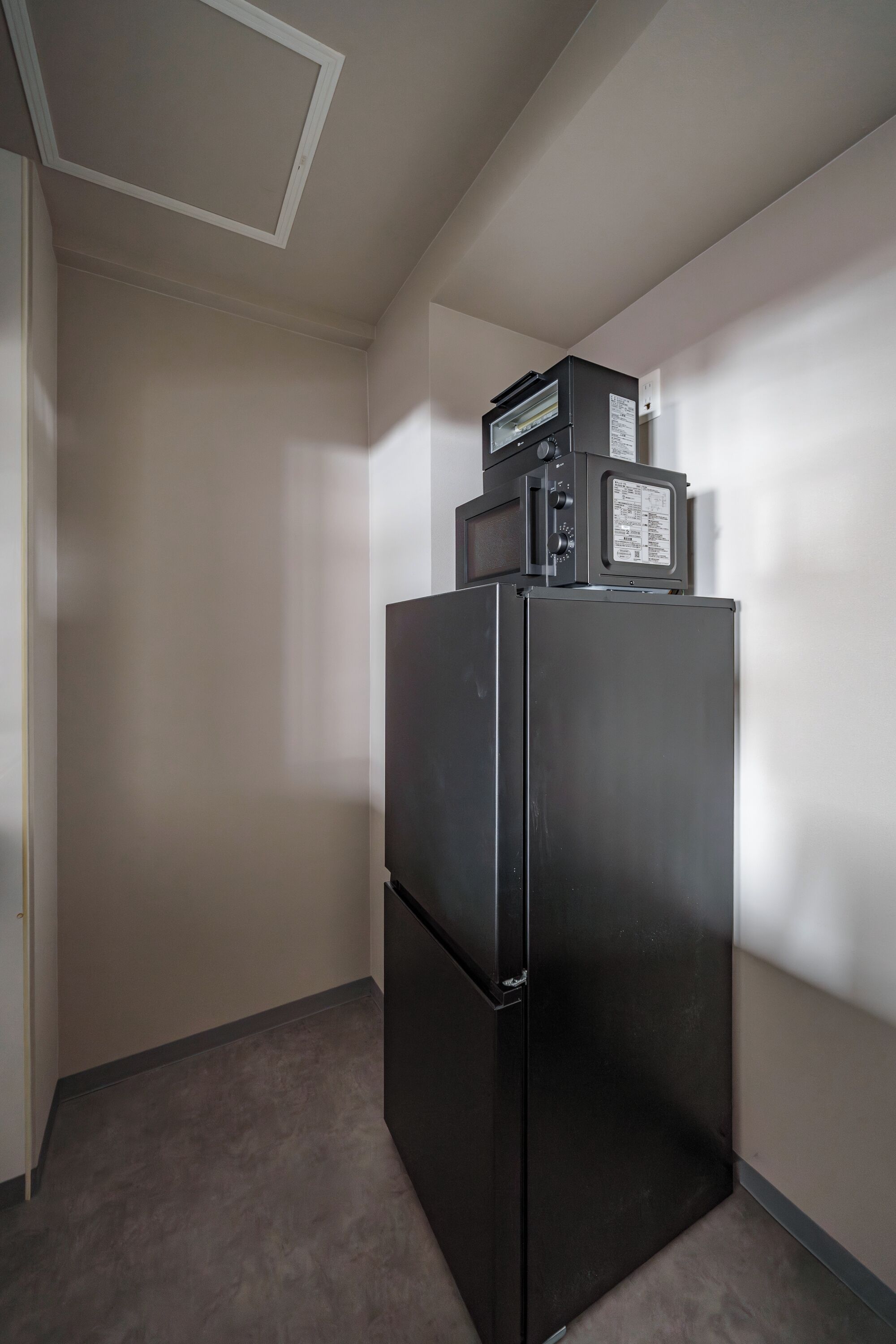 A small, dark-colored refrigerator with a microwave on top, located in a corner of a room with plain walls and a carpeted floor.