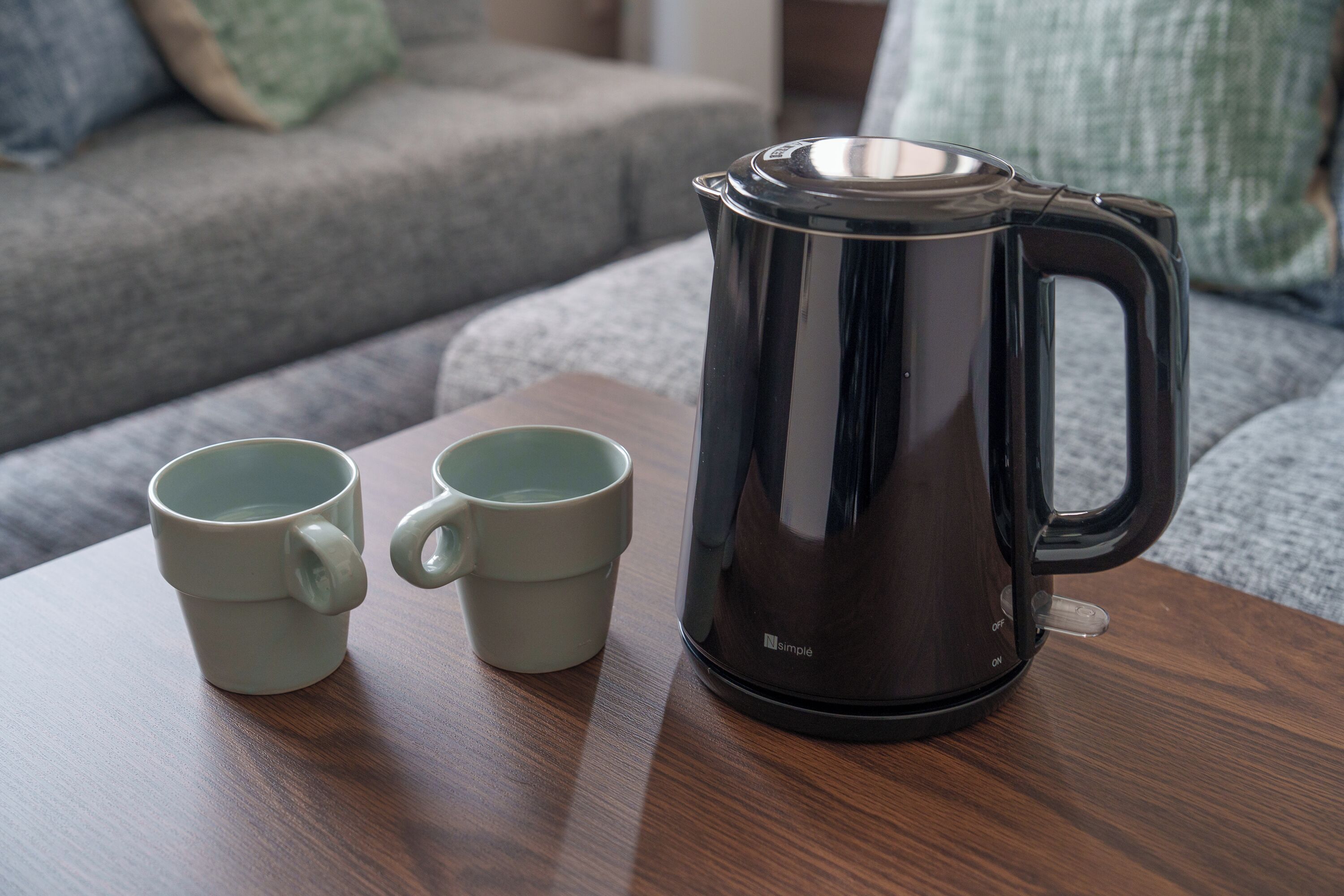 A coffee maker and two mugs on a wooden table in a living room.