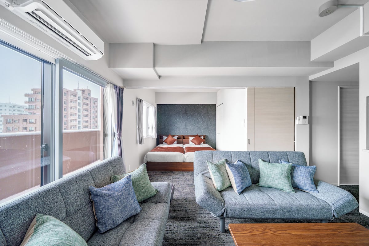 A modern hotel room with a large window, a bed with a headboard, a grey sofa set, and a wooden coffee table.