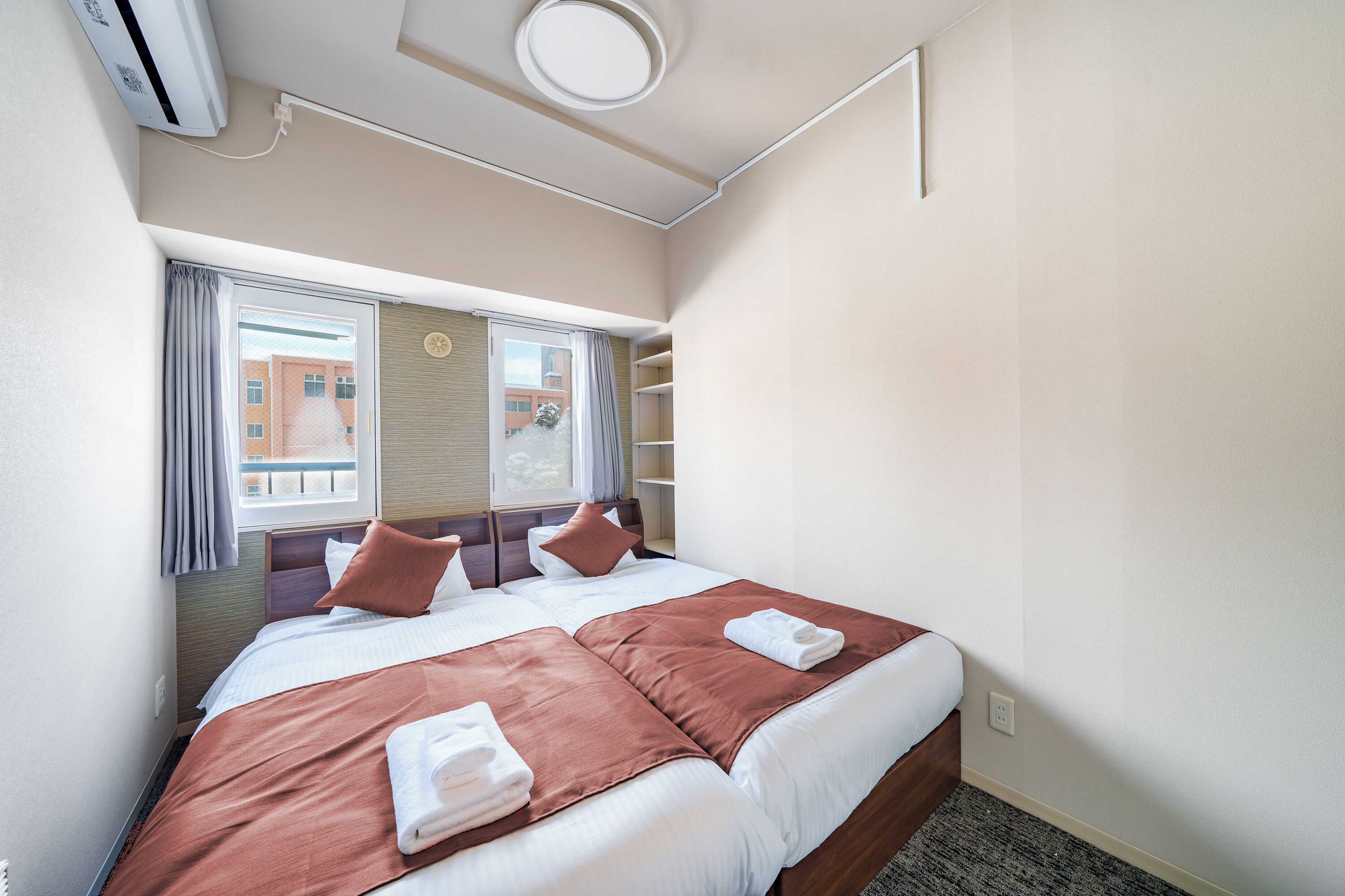 A hotel room with two beds, a window with curtains, a wall-mounted air conditioner, and a view of buildings outside.