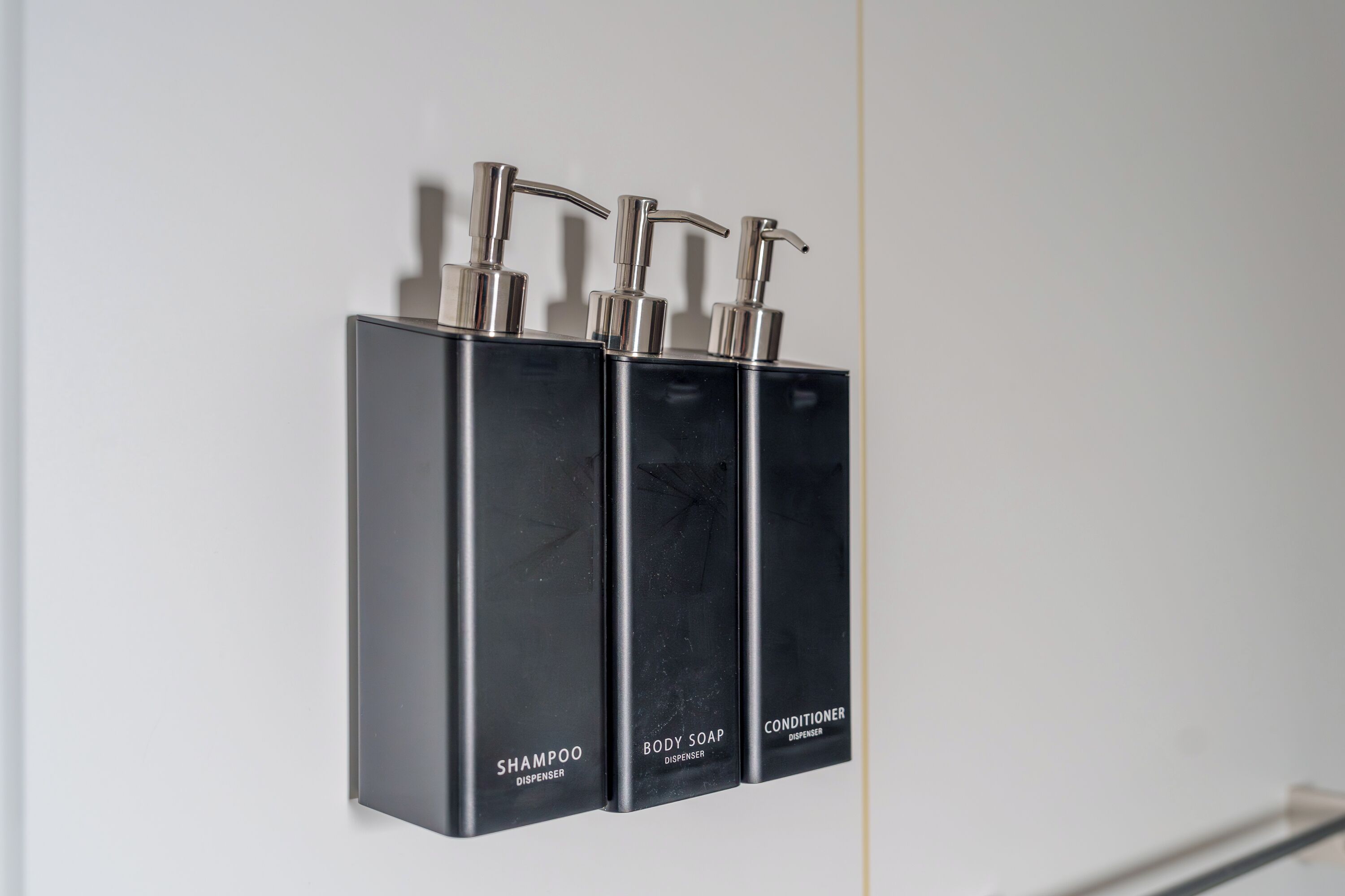 Three black dispensers labeled 'Shampoo', 'Body Soap', and 'Conditioner' mounted on a wall.