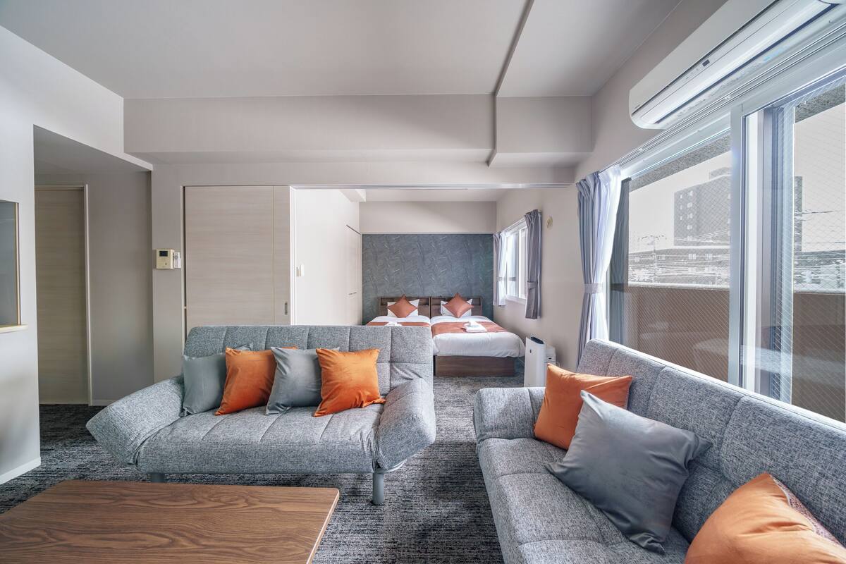 A modern hotel room with a grey sofa set, a wooden coffee table, and a bed with orange and grey pillows.