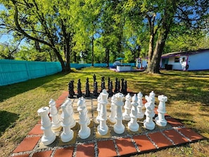 A large outdoor chessboard set up on a paved area with trees and a building in the background.
