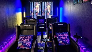 Une pièce comprenant quatre fauteuils noirs inclinables portant l’inscription « Boss N », un panneau néon indiquant « Soirée cinéma » et un plancher en bois.