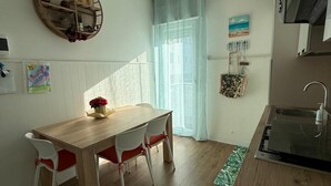 Eine kleine Küche mit Holztisch und -stühlen, einer Pendelleuchte, einem runden Regal mit Dekoration und einem Fenster mit transparenten Vorhängen.