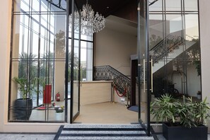 L’entrée moderne d’un hôtel, avec une façade en verre, un lustre, un escalier orné de rampes travaillées et des plantes en pot.
