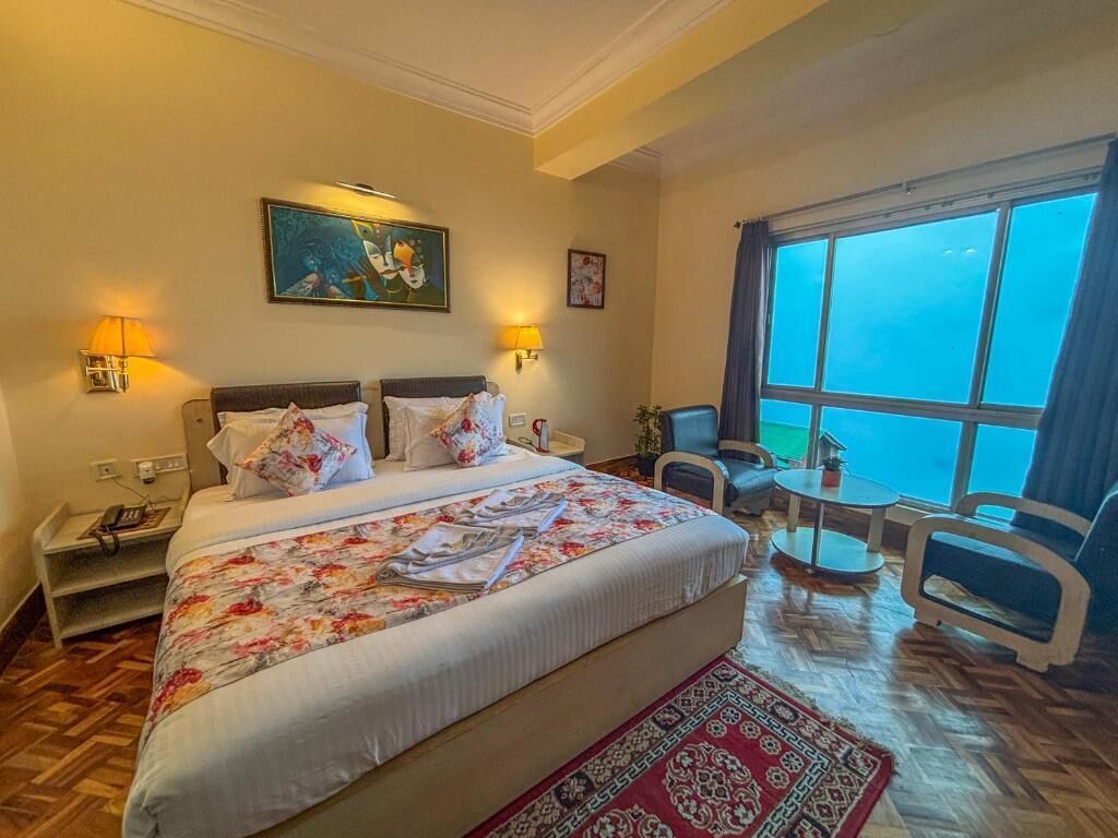 Habitación de hotel con una cama grande, dos lámparas de noche, una mesita con una silla y un ventanal con vistas.
