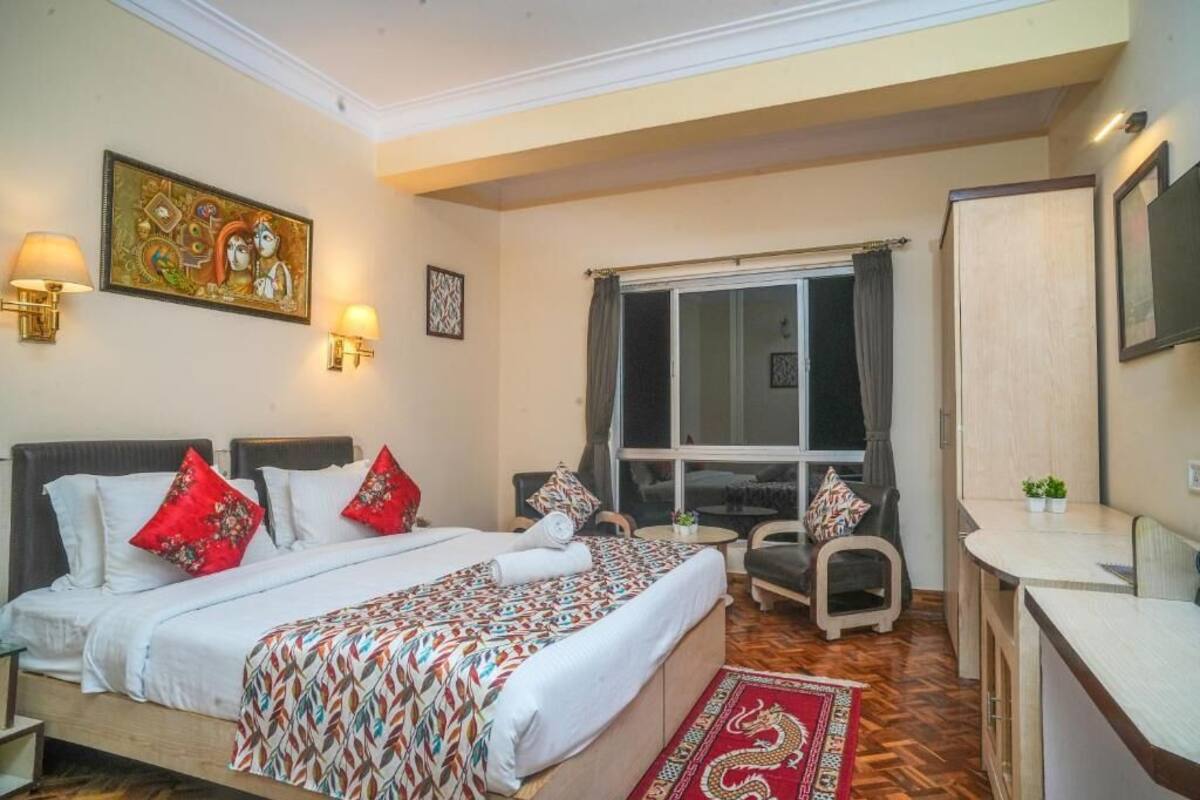 A hotel room with a bed, bedside tables, a desk, a chair, a window with curtains, and a painting on the wall.