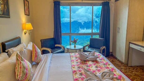 Ein Hotelzimmer mit einem Bett, einem Schreibtisch, einem Stuhl, einem kleinen Tisch mit einer Vase und Meerblick durch große Fenster.