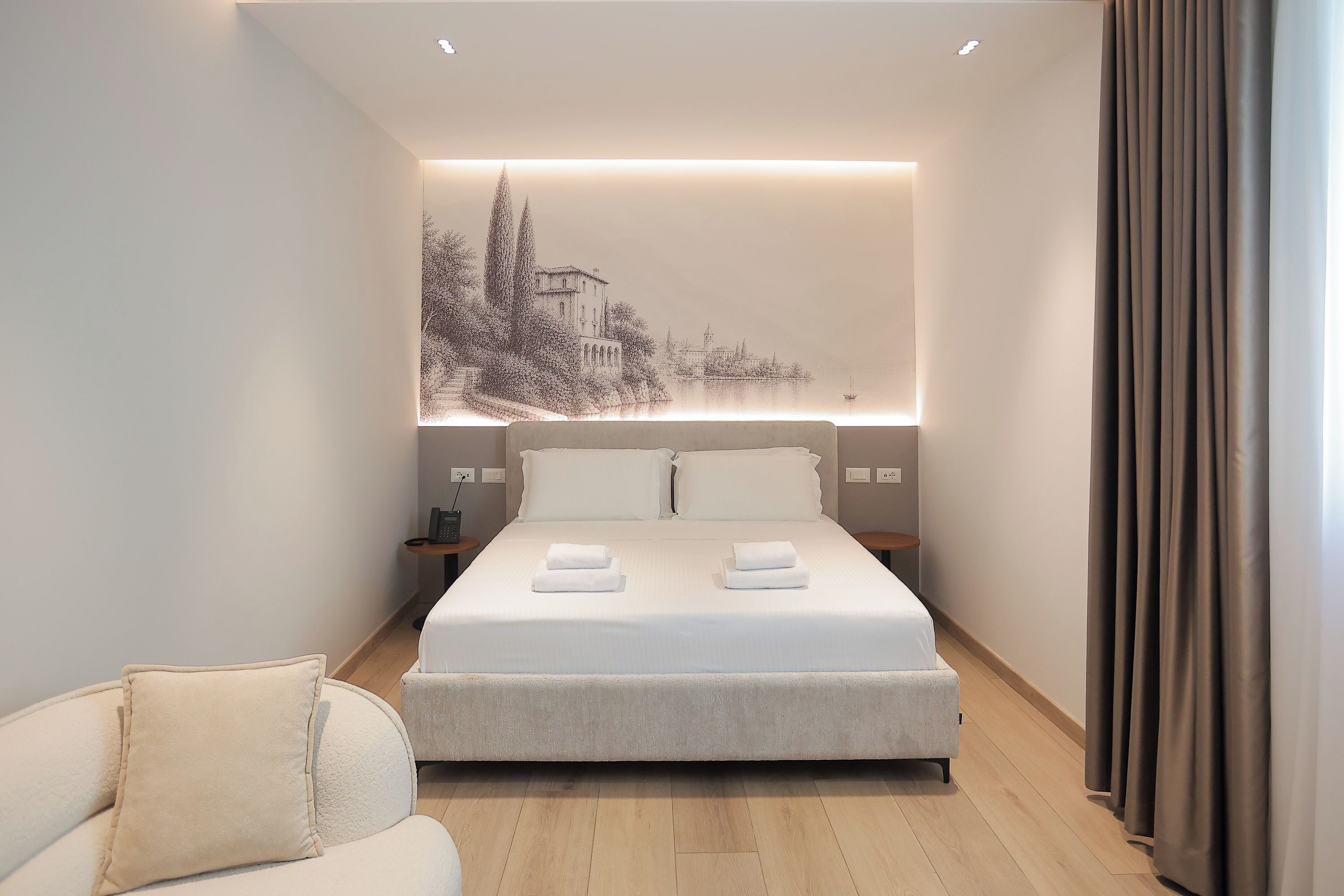 A modern hotel room with a large bed, bedside tables, a nightstand, a sofa, and a wall-mounted artwork.