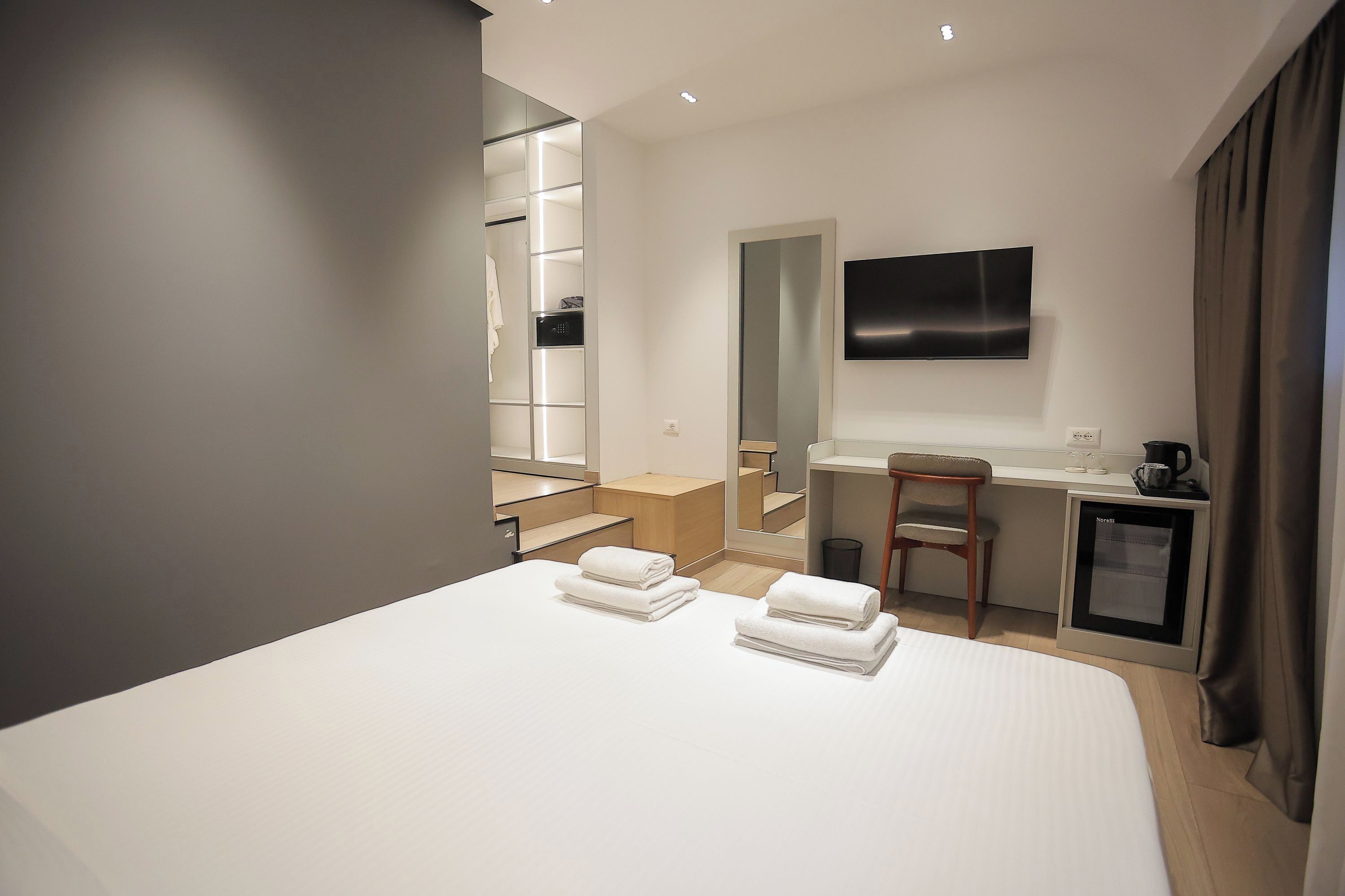 A modern hotel room with a large bed, a desk, a chair, a TV, and a walk-in closet.