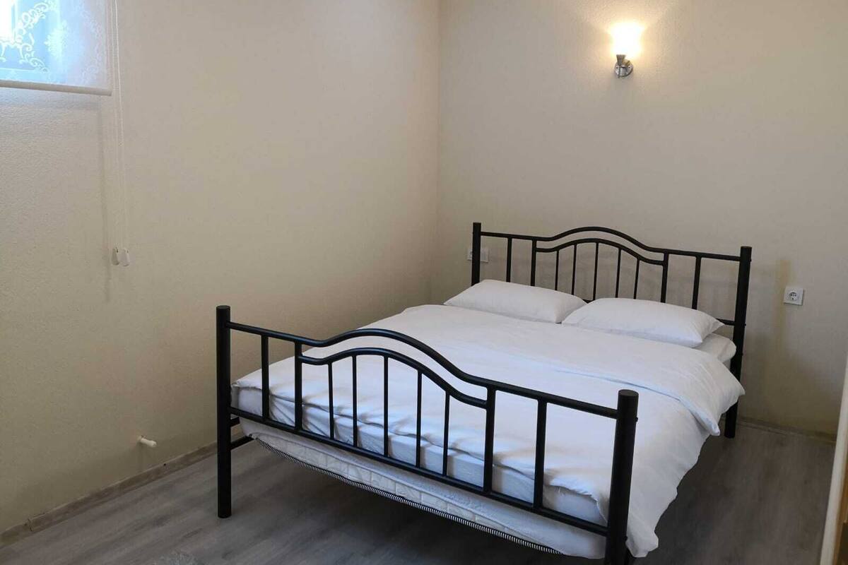 Una cama individual con estructura metálica negra y ropa de cama blanca en una habitación con paredes color beige y un interruptor de luz en la pared.