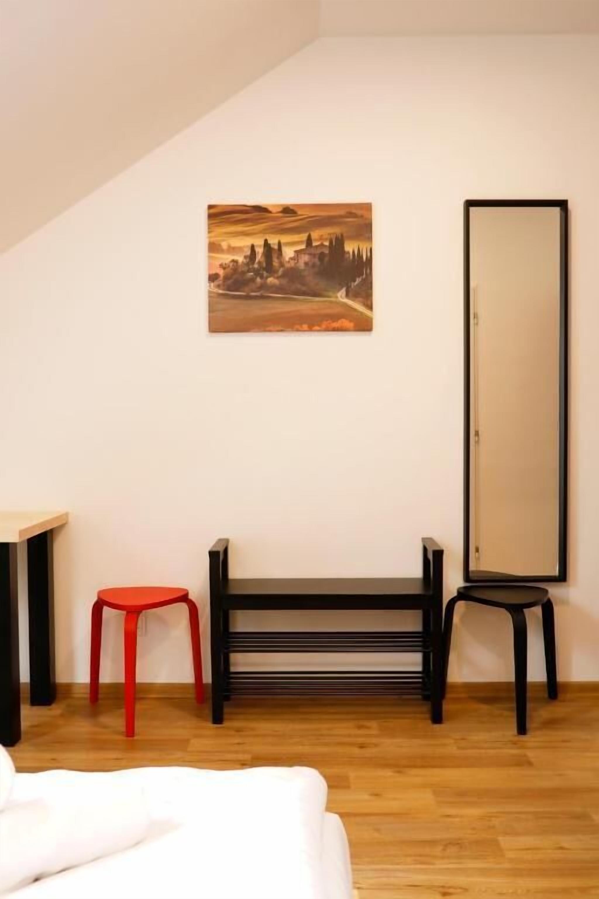 A room with a wooden floor, a bed, a red stool, a black bench, a large mirror, and a painting on the wall.