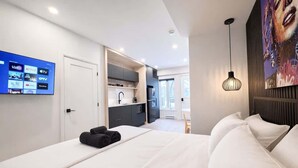 Et moderne hotelværelse med seng, fjernsyn på væggen der viser streamingtjenester, et spiseområde synligt gennem en åben dør, og en pendel over sengen.