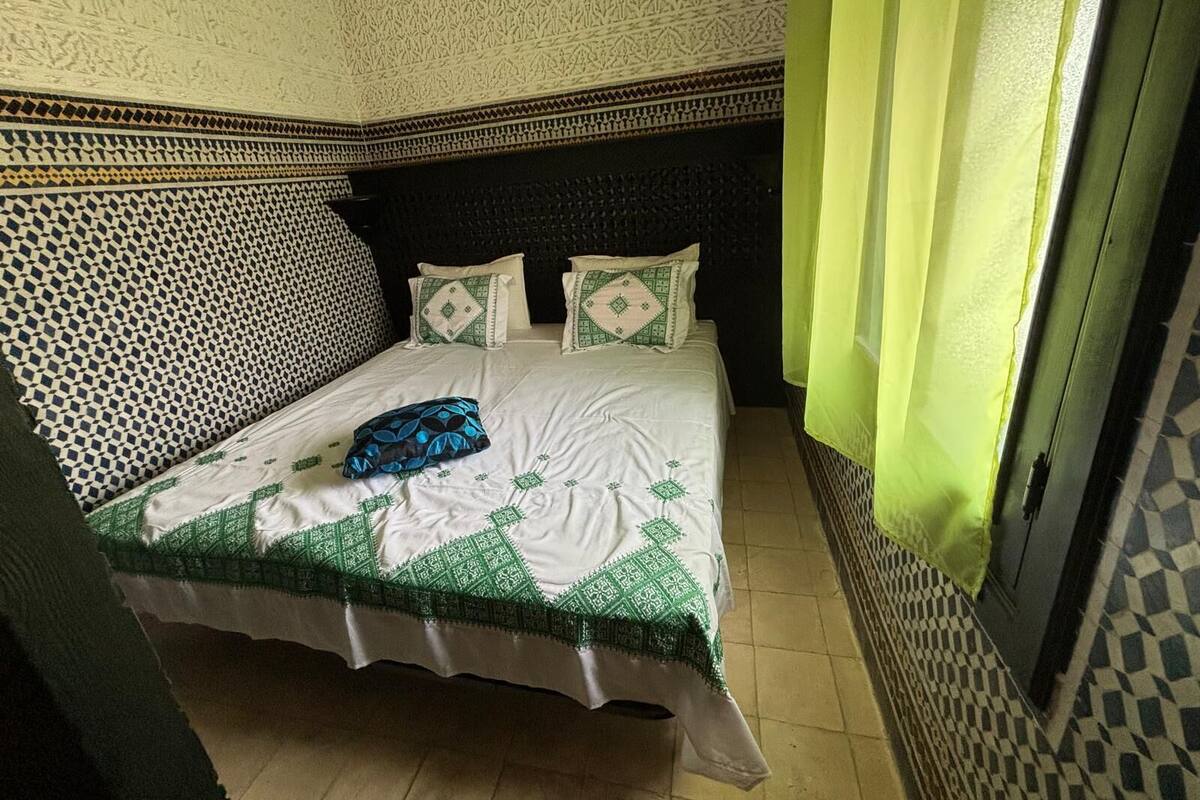 Une pièce avec un mur orné de motifs, un lit recouvert d’une couvre-lit verte et blanche, et une tête de lit noire.
