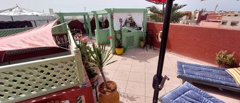 Una terraza en la azotea con un paraguas rojo, decoración verde y blanca, y un área de estar con cojines azules.