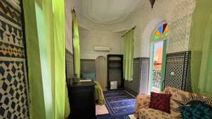 A room with patterned walls, a bed with patterned bedding, a desk with a lamp, and a window with a view. - Riad Zahrat Al Fajr (El Jadida)