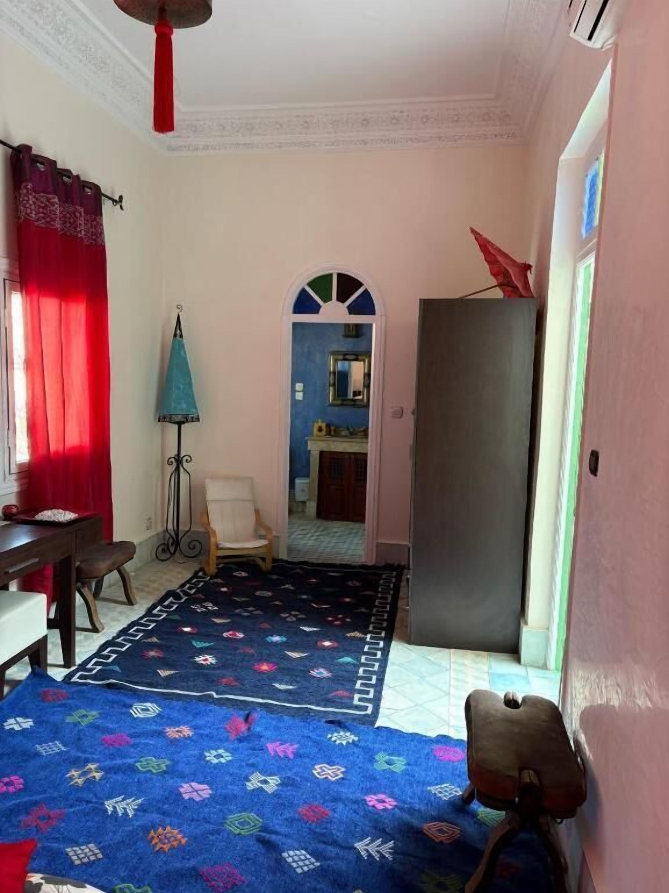 A room with a patterned carpet, a dining table, a chair, a red curtain, and a door leading to another room.