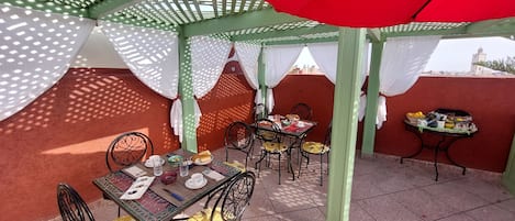 An outdoor patio with a table set for dining, covered by a red umbrella, and surrounded by chairs with patterned cushions.