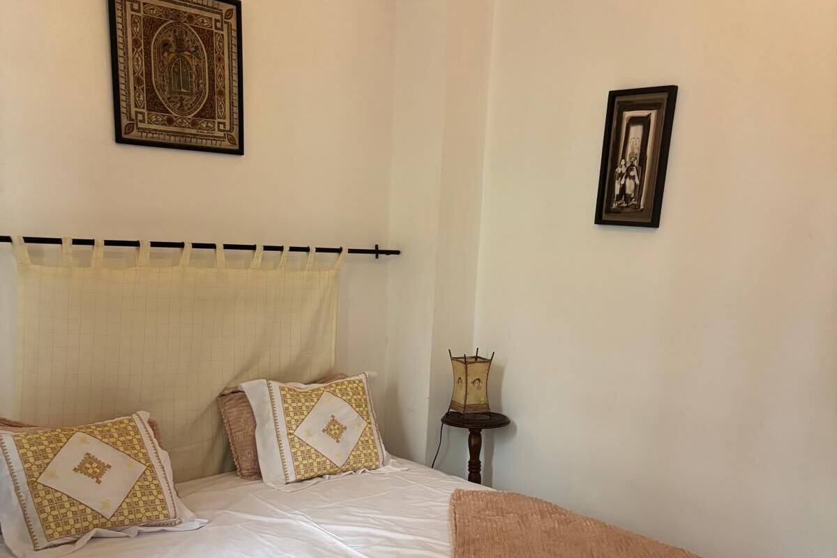 Un lit bien fait, avec une literie à motifs, une petite table de chevet sur laquelle se trouve une lampe, et un tableau encadré accroché au mur.