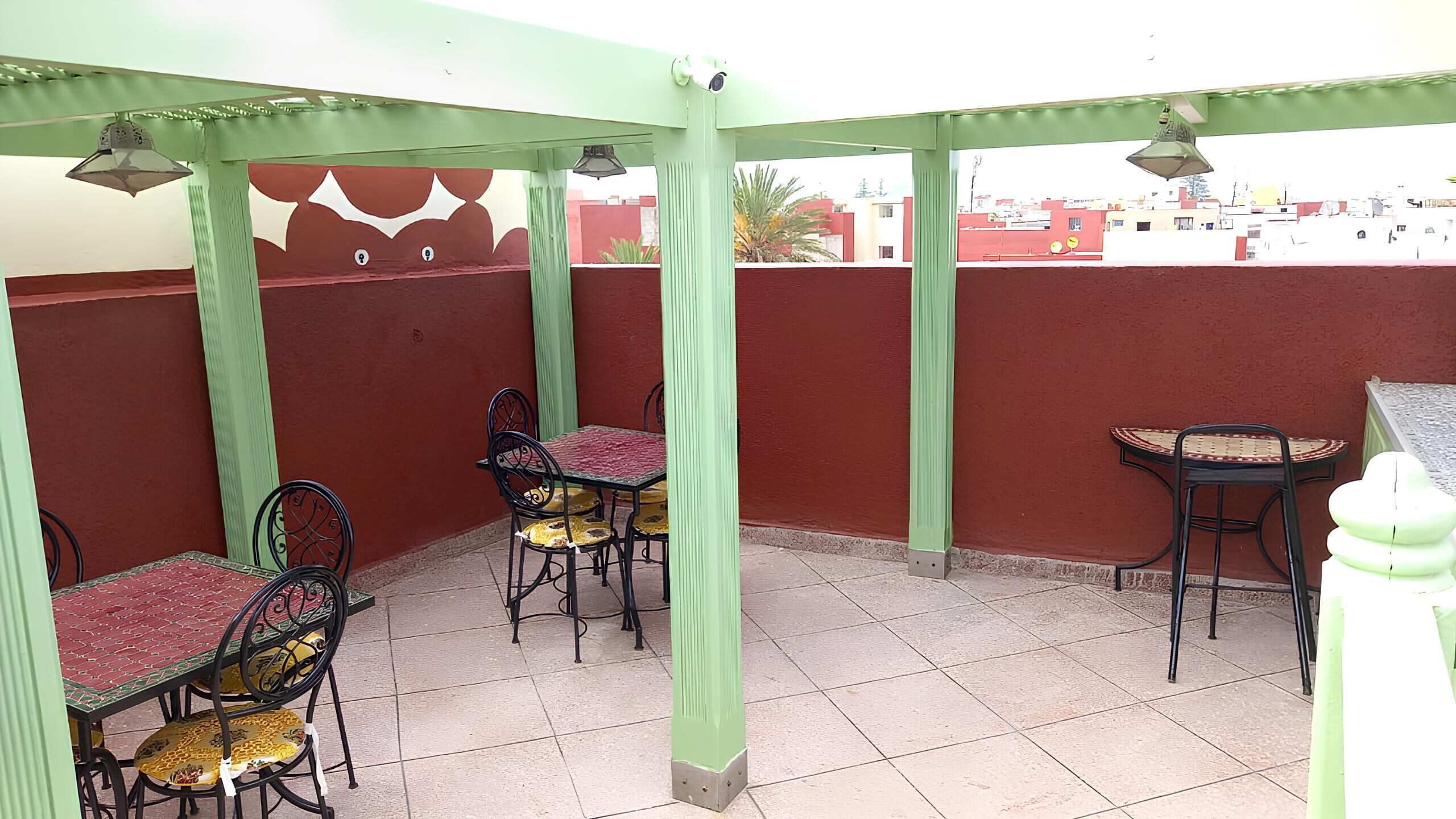 Un espacio de estar al aire libre cubierto con mesas y sillas, pilares verdes y vistas a edificios al fondo.