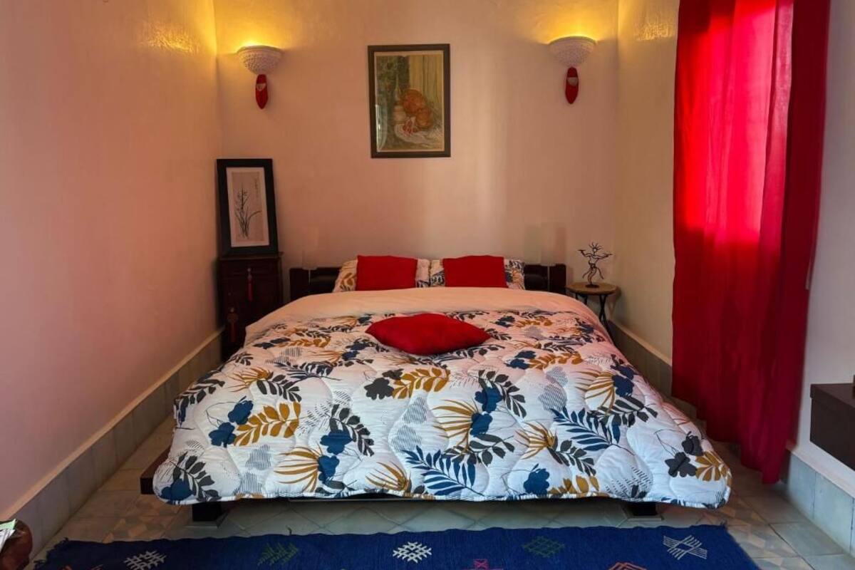 Une chambre à coucher avec une couvre-lit à motifs floraux, des coussins rouges, un tableau encadré au mur et un rideau rouge.