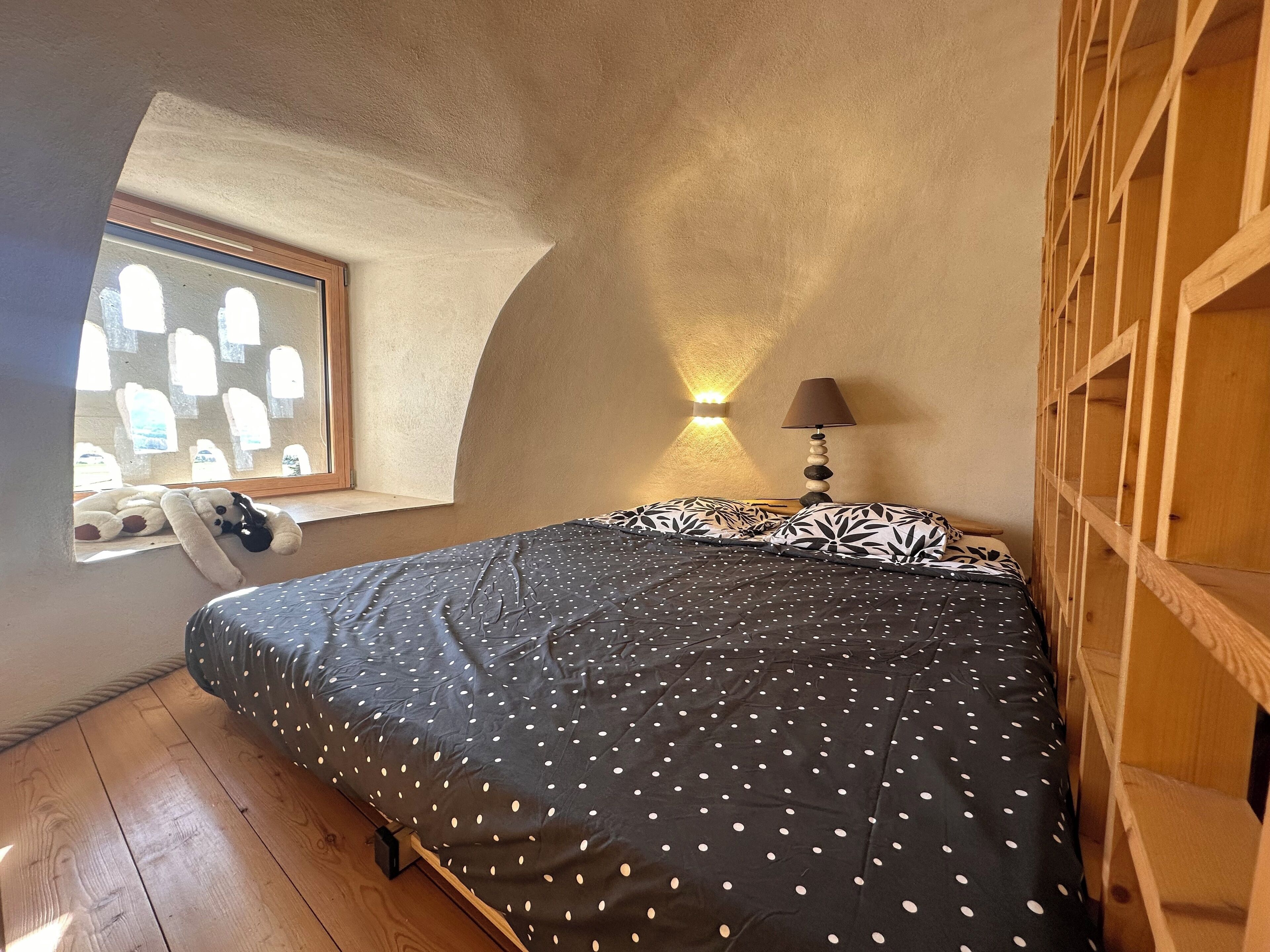 Un dormitorio con cama, ventana, lámpara y estante de madera.