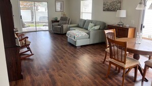 A spacious living room with a wooden floor, a grey sofa, a wooden dining table with chairs, and a ceiling fan. - The Villages FL. Courtyard, Villa House, Golf Cart, Dogs Welcome, Fenced Yard. (The Villages)