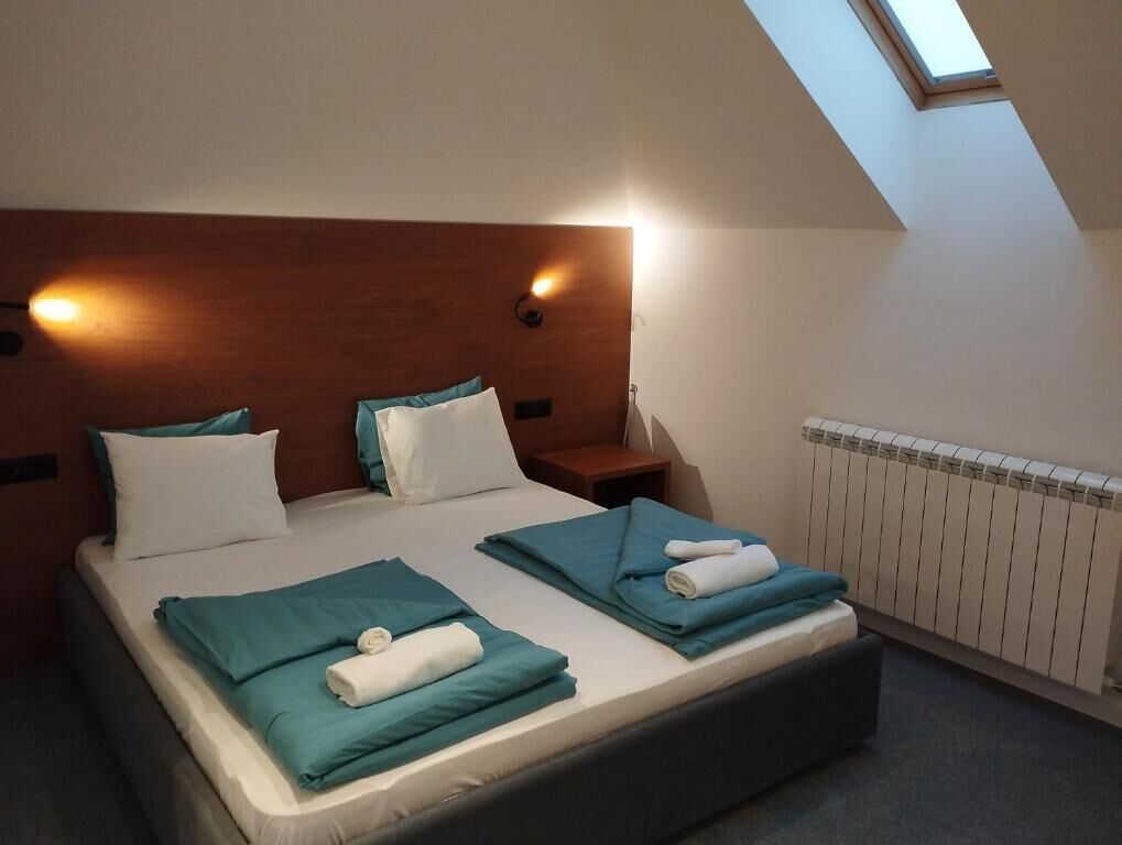 Un dormitorio con techo inclinado, cabecera de madera, dos camas con ropa de cama color turquesa, una mesita de noche y un tragaluz.