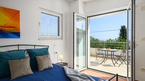 Un dormitorio con cama, almohadas, una mesita de noche, una lámpara y vistas a un balcón exterior con mesa y sillas.