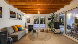 Una sala de estar moderna con techo de madera, un sofá con cojines coloridos, una mesa de centro, un televisor y grandes puertas de vidrio correderas que dan a un patio exterior.