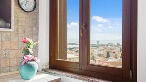 Ein Fenster mit Blick auf eine Stadtlandschaft, eine Uhr an der Wand und eine Vase mit Blumen auf einem Fensterbrett.