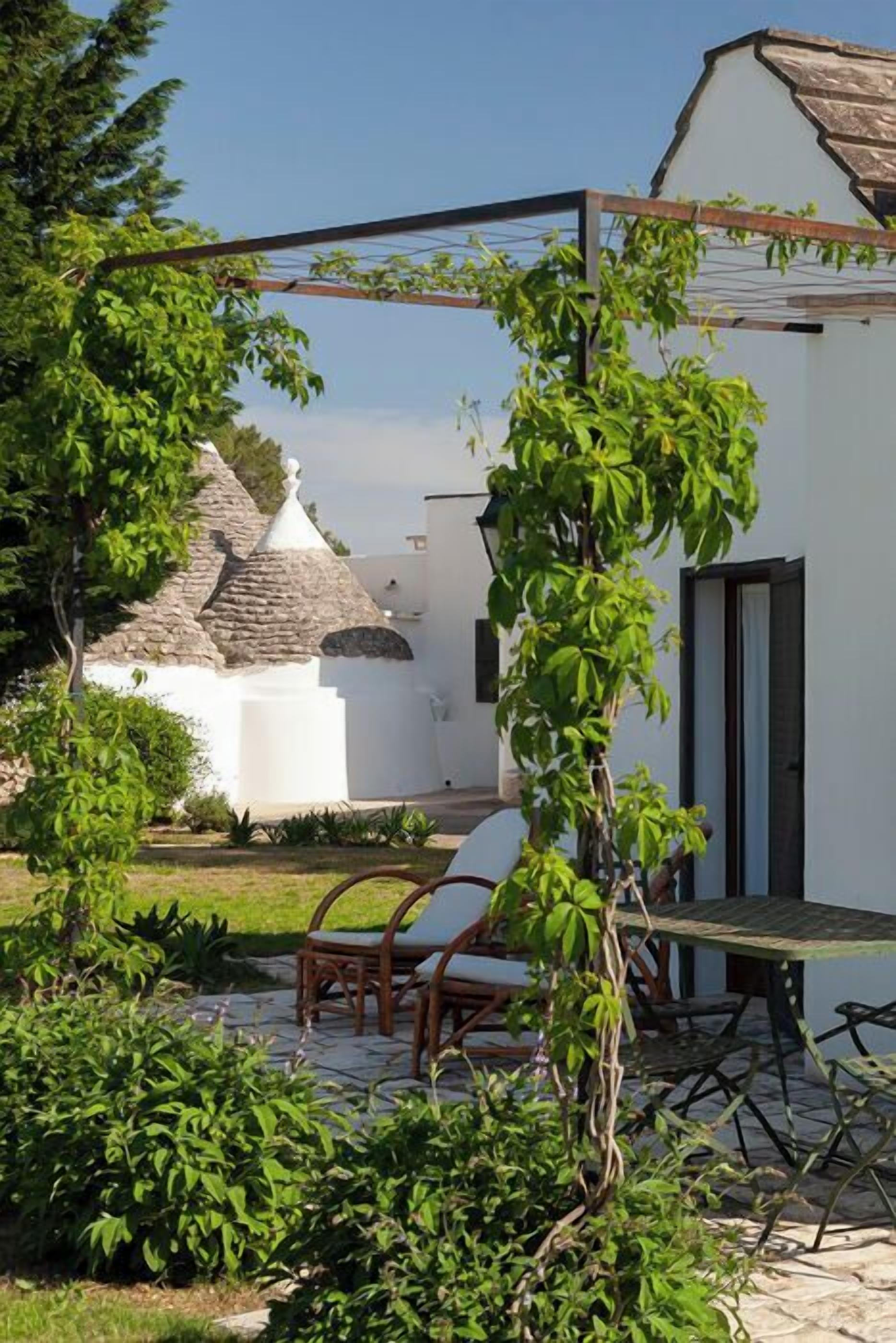 A patio with a table and chairs, a white building with a thatched roof, and a garden with various plants.