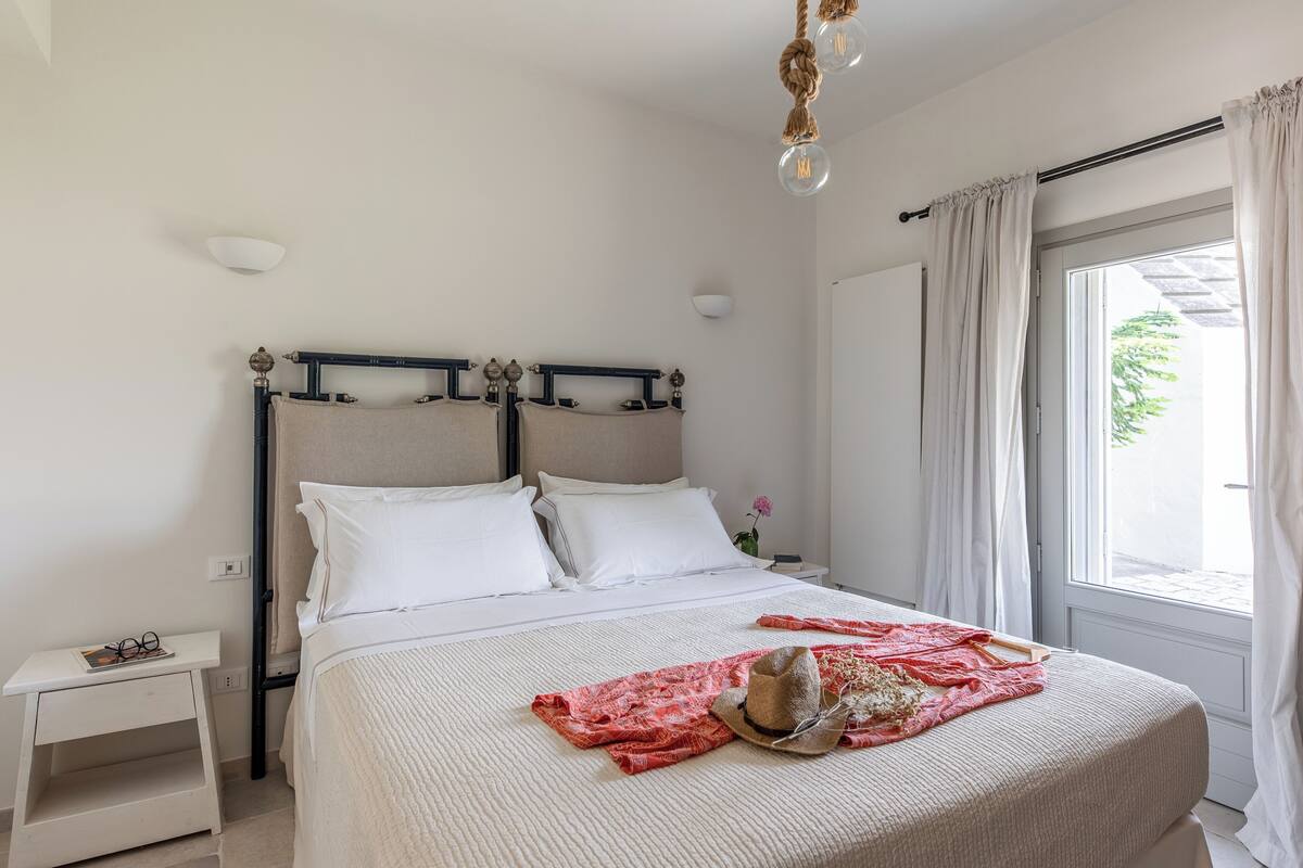 A neatly made bed with a beige blanket and a red cloth, a small bedside table with glasses, and a window with curtains.
