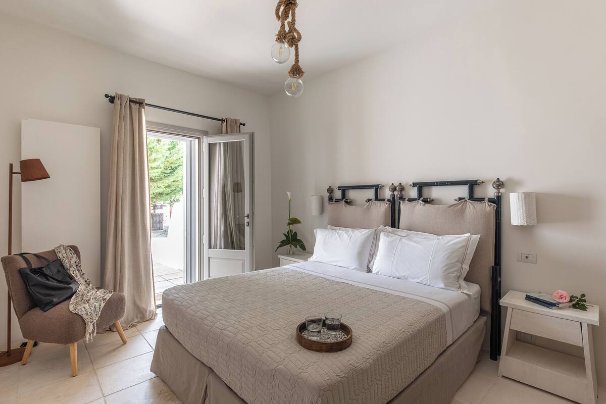 A bedroom with a bed, bedside table, lamp, chair, and a view of the outdoors.