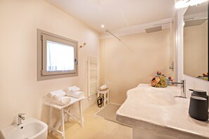 Un baño con lavamanos, bidé y ventana. Hay toallas sobre una mesita y un objeto decorativo en el mostrador.