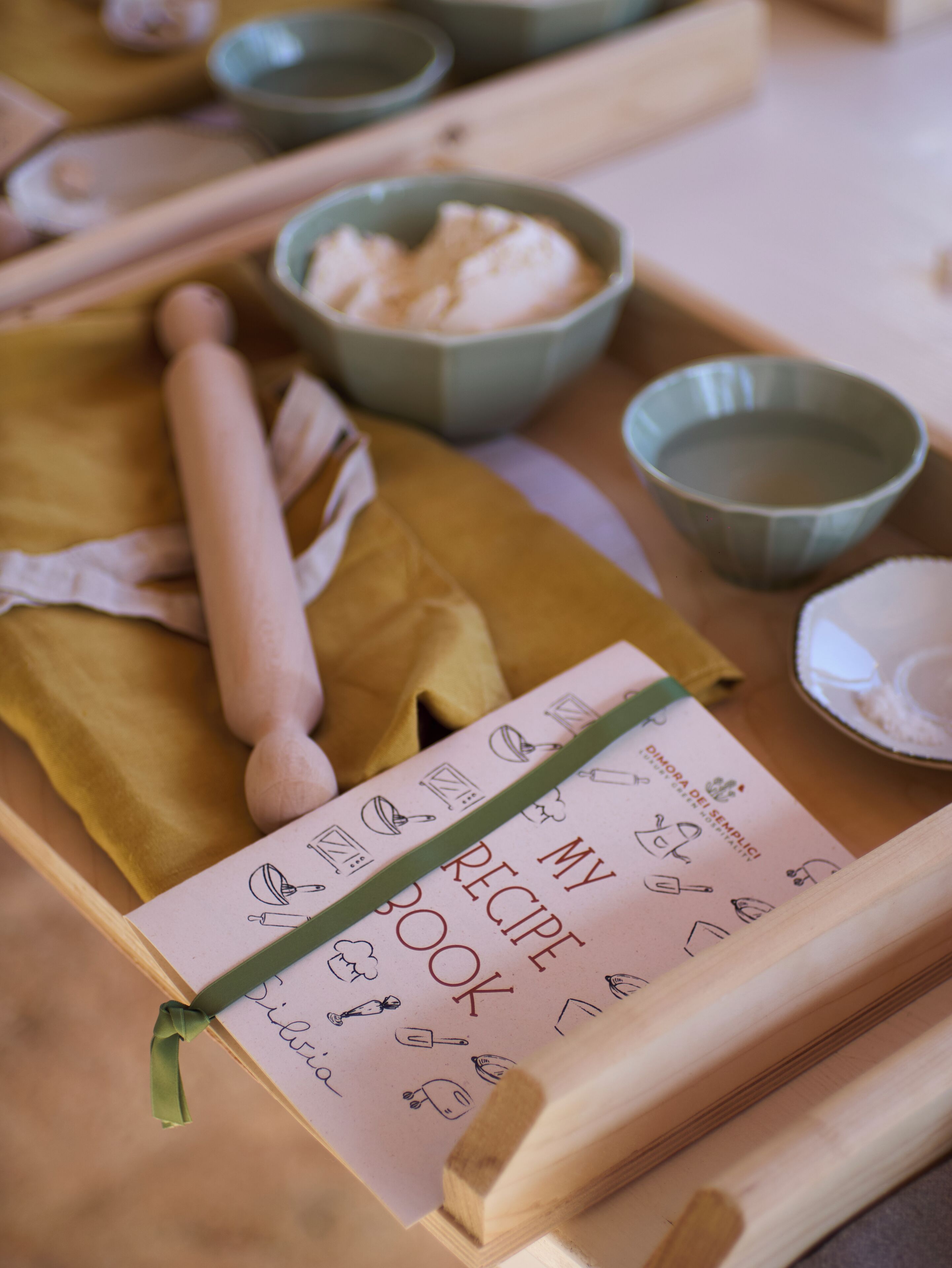 Un safata de fusta amb un llibre de receptes, un rodillo i cuissos d'ingredients.