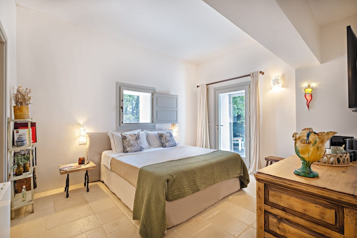 A bedroom with a bed, bedside table, lamp, and a view of the outdoors.