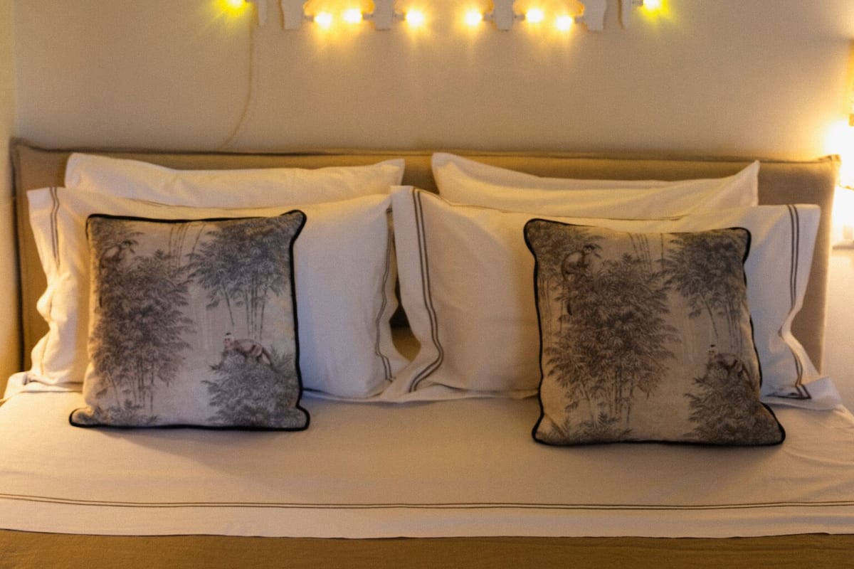 A bed with white bedding and two decorative pillows, a wall-mounted light fixture with multiple bulbs, and a wooden headboard.