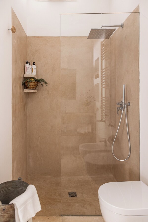 A glass-enclosed shower with a handheld showerhead, a white toilet, and a shelf with toiletries. - Dimora dei Semplici (Ostuni)