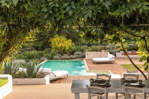 Un área junto a la piscina con cubierta de madera, un espacio de estar exterior blanco y diversas plantas y arbustos.