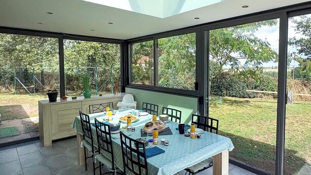 Un espace repas avec une table dressée pour quatre personnes, entouré de grandes fenêtres et éclairé par une lucarne.
