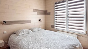 Une chambre à coucher avec un lit, un luminaire suspendu, une fenêtre avec des stores et un mur orné d’un motif géométrique.