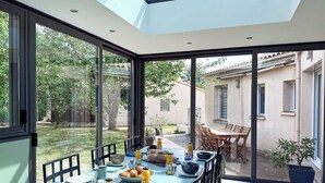 Un coin repas avec une table dressée pour quatre personnes, de grandes fenêtres et une vue sur le jardin et les bâtiments voisins.