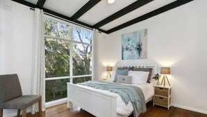 Un dormitorio con una cama blanca, una silla gris, un ventanal grande y un cuadro en la pared.