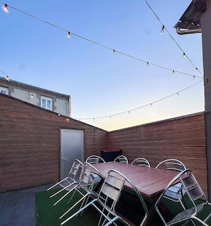 An outdoor patio with a table and chairs, string lights, and a wooden building in the background. - Tranquiloue - Gite 15 personnes - 10min Puy du Fou (Saint-Mars-la-Réorthe)