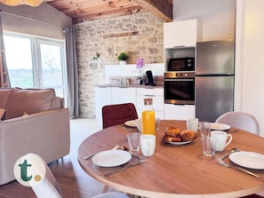 Un coin repas avec une table en bois dressée pour le petit-déjeuner, un mur en pierre, une cuisine équipée d’appareils modernes et une vue par la fenêtre.