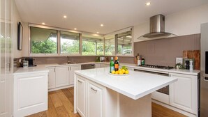 A modern kitchen with white cabinetry, a central island with fruit, and a large window offering a view of greenery. - Yera Coastal Homestead with Tennis Court & Spa (Rye)