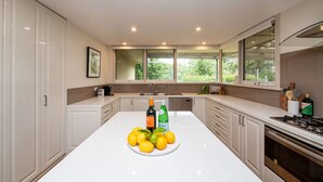 A modern kitchen with white cabinetry, a central island with fruit and a bottle, and a view of greenery outside. - Yera Coastal Homestead with Tennis Court & Spa (Rye)